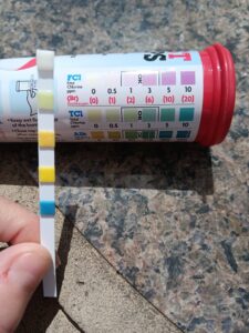 A hand holding a pool water test strip next to a color chart, demonstrating water chemistry testing by Fox Pools of Evansville Inc in Evansville, IN