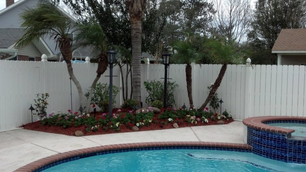 A beautifully landscaped poolside flower bed with palm trees and a white fence by Fresh Cuts Nola Lawn Service in New Orleans, LA.