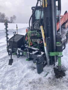 A post driver attachment on a Kubota skid steer drilling a hole in snowy ground for fence installation by Doolan Fence in Montpelier, VT.