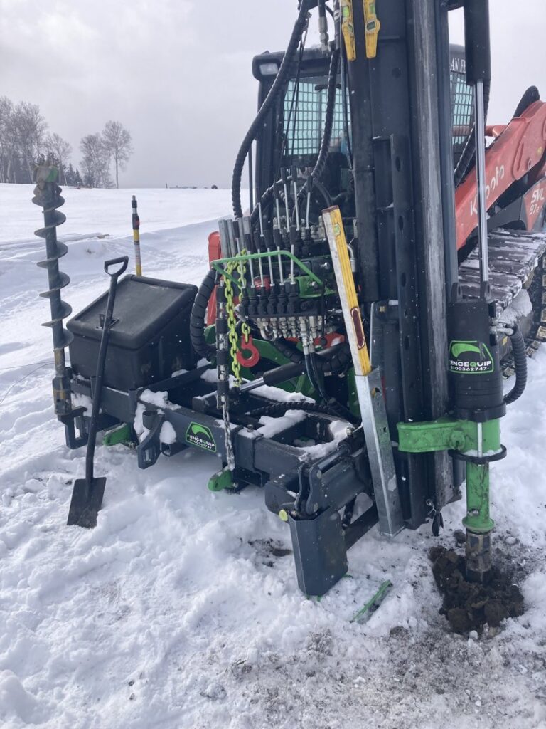 A post driver attachment on a Kubota skid steer drilling a hole in snowy ground for fence installation by Doolan Fence in Montpelier, VT.