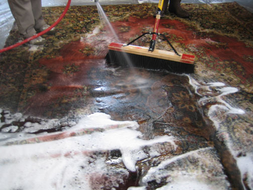 Professional rug cleaning in progress with water and a large brush at Heriz Rugs, Art, & Interior Design Services in Santa Fe, NM.