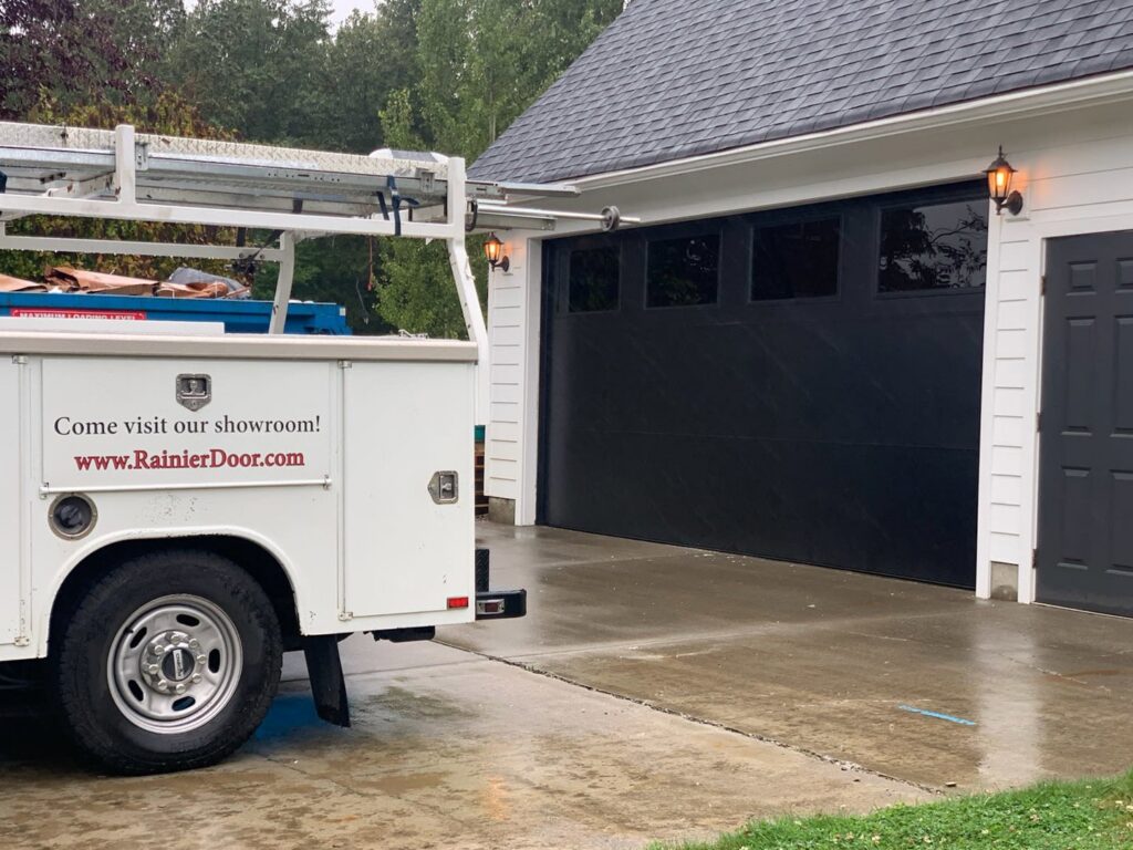 A Rainier Garage Door service truck parked at a residential job site with a new black garage door in Bellevue, WA.