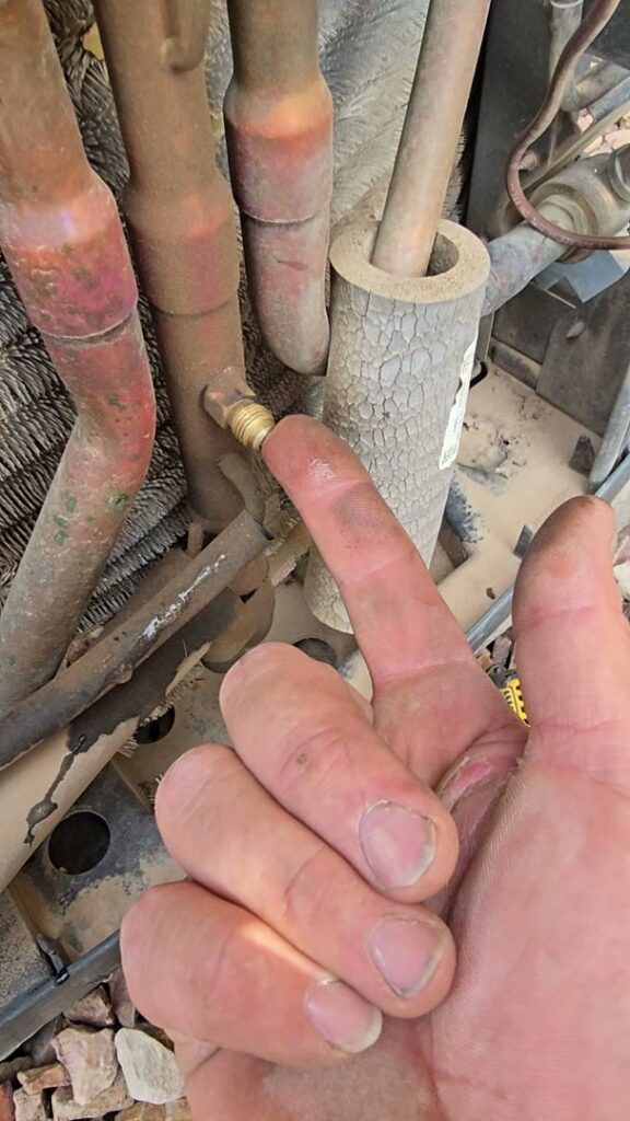 A hand pointing to a brass fitting on a copper refrigerant line of an outdoor AC unit by Quantum Cooling & Heating in Mesa, AZ.