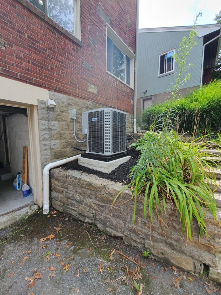 A residential air conditioning unit installed next to a house with landscaping by Bouvy Heating & Air Conditioning Services in Pittsburgh, PA.