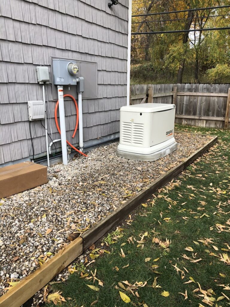 A Generac generator installed on a gravel bed next to a residential building with electrical connections by Great Lakes Power Generation in Williamsburg, MI.