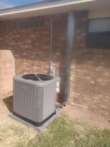 A Rheem outdoor HVAC condenser unit installed next to a brick house by Raytech Refrigeration, Heating & Cooling in Alamogordo, NM.