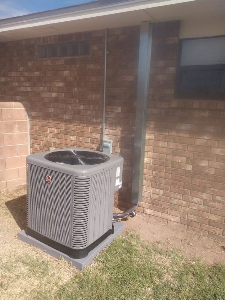 A Rheem outdoor HVAC condenser unit installed next to a brick house by Raytech Refrigeration, Heating & Cooling in Alamogordo, NM.