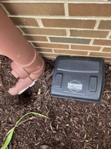 A black rodent bait station with the company logo placed near a house foundation on mulch by Modern Solutions Pest Control in Rockford, IL.