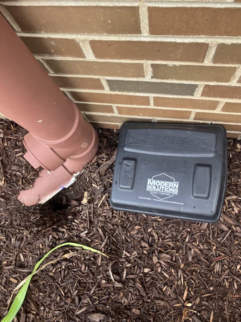 A black rodent bait station with the company logo placed near a house foundation on mulch by Modern Solutions Pest Control in Rockford, IL.