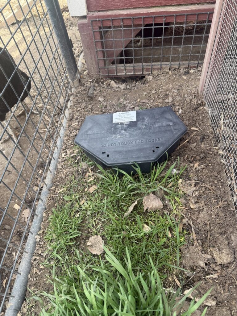 A black rodent bait station placed on the ground near a fence, indicating pest control activity by Peak Pest Control, Ogden, UT.