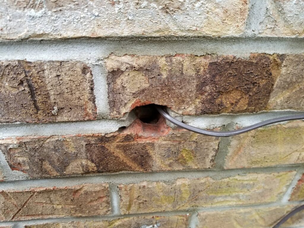 A small hole in a brick wall with a wire, potentially a rodent or pest entry point, addressed by Spectrum Pest Control Eco-Tech LLC in Kenosha, WI.