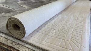 A rolled light-colored rug displayed on top of other rugs, showcasing inventory at Makymo in Commerce, CA
