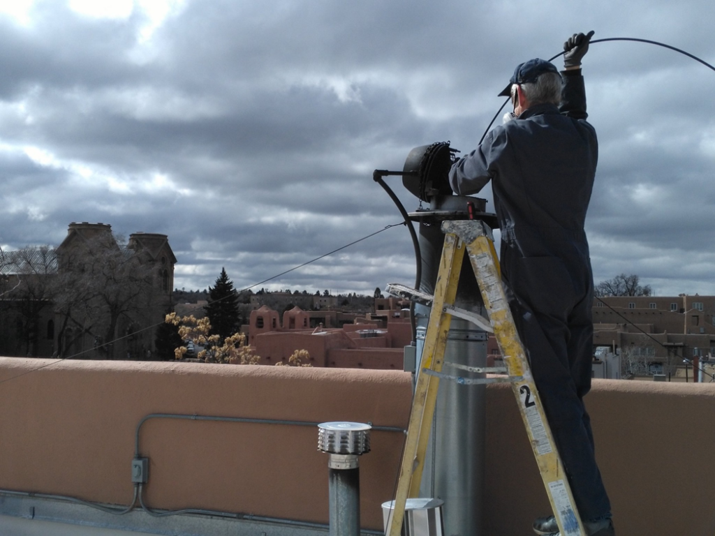 A K-West NM technician on a ladder performing rooftop chimney cleaning with the Santa Fe, NM skyline in the background.