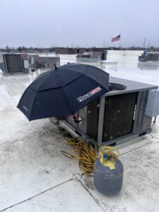 A rooftop HVAC unit being serviced in snowy weather with an umbrella and refrigerant tank by KCC Mechanical, LLC in Sandy, UT.