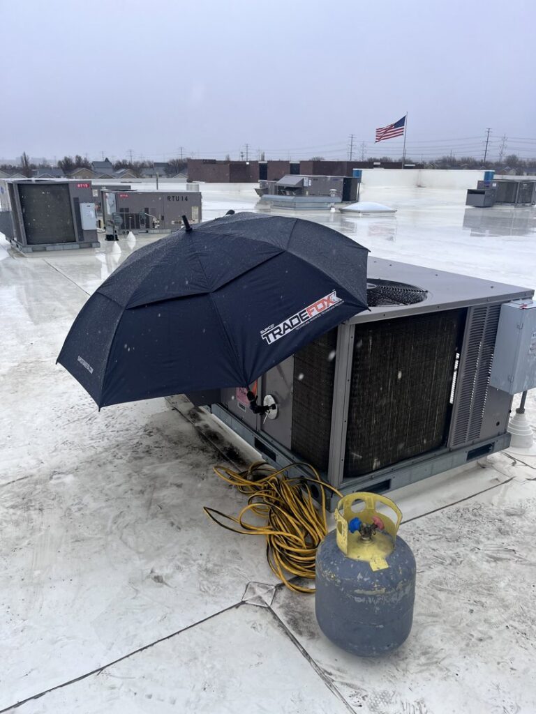 A rooftop HVAC unit being serviced in snowy weather with an umbrella and refrigerant tank by KCC Mechanical, LLC in Sandy, UT.