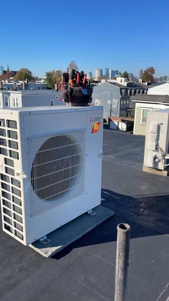 A Mitsubishi mini-split outdoor unit on a rooftop with the Boston skyline in the background, installed by EML Mechanical in Boston, MA.