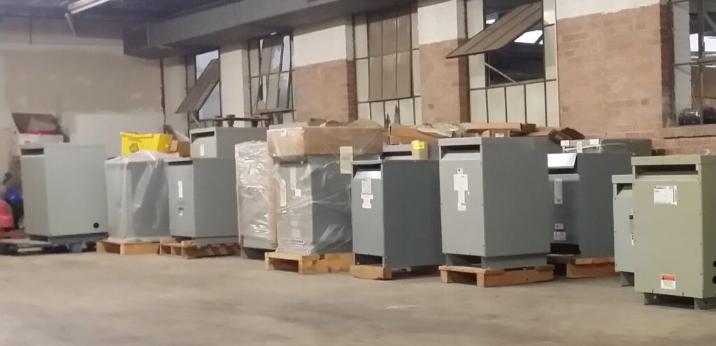 A row of large electrical transformers or enclosures stored on pallets, available from Basic Supply in Lafayette, LA.