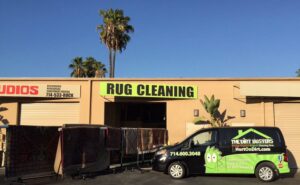 Rugs hanging on racks outside a building with a 'RUG CLEANING' sign, next to a Dirt Busters, Inc. service van in Anaheim, CA.