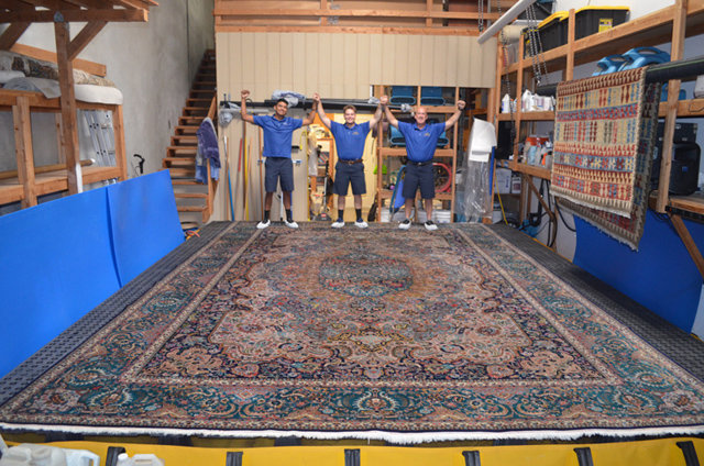 The Rug Renew team standing proudly behind a large, beautifully cleaned rug in their facility in Spring Valley, CA.