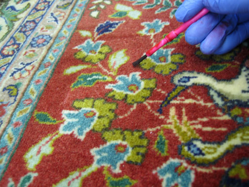 A gloved hand meticulously repairing or restoring a patterned rug at Heriz Rugs, Art, & Interior Design Services in Santa Fe, NM.