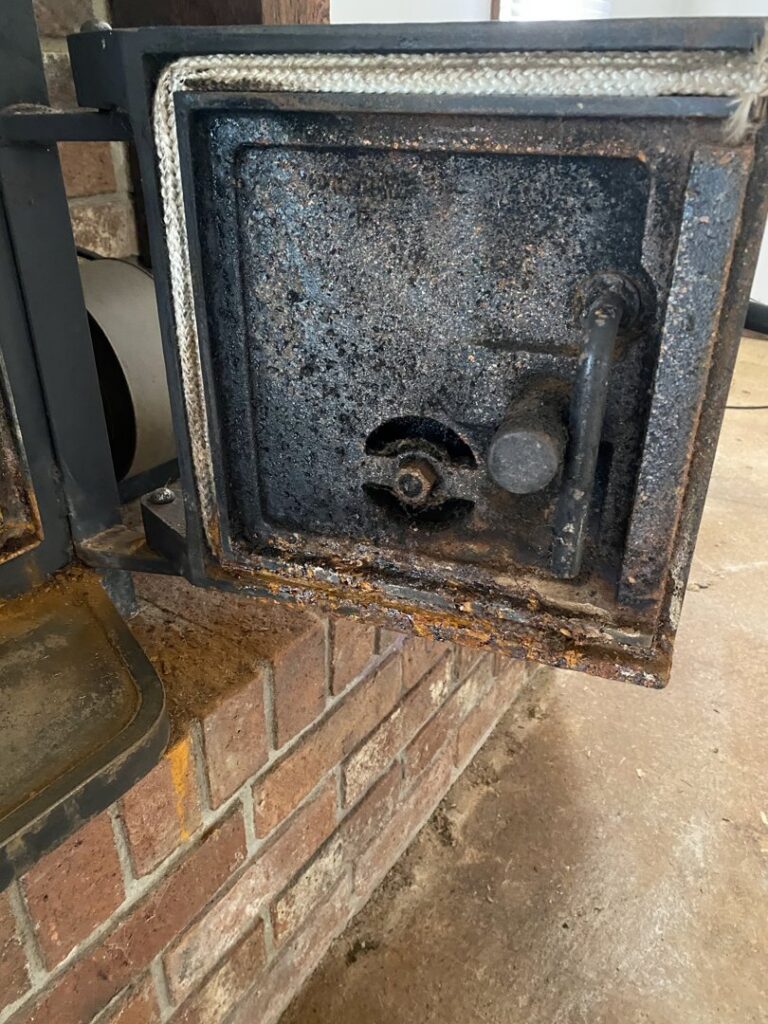 A close-up of a rusty and dirty fireplace insert door, indicating a need for maintenance by Chimney Cricket Cleaning Service in Marshfield, MO