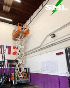 A scissor lift extended to reach electrical conduit on a high wall, indicating an installation or repair job by SML Electrical in Pearl City, HI.