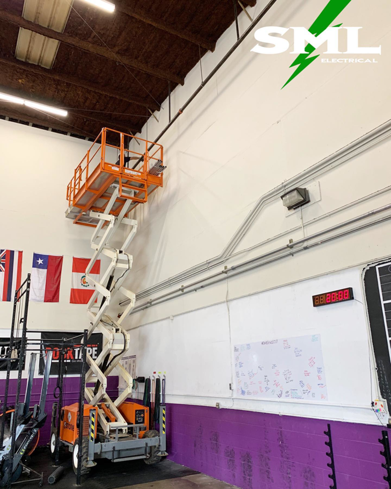A scissor lift extended to reach electrical conduit on a high wall, indicating an installation or repair job by SML Electrical in Pearl City, HI.