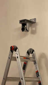 A security camera mounted on a wall with a ladder, indicating installation work by Gksquad in Columbia, SC.