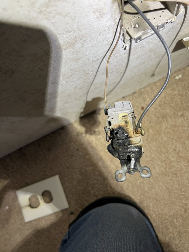 A severely burnt electrical outlet removed from the wall, with a new cover nearby, indicating a repair by KNK Electric in Pittsburgh, PA.