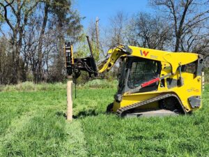 A skid steer with a post driver attachment installing a wooden fence post for H&S Fencing & Supply in Linden, WI.