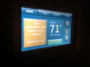 A smart thermostat displaying a message for humidifier pad replacement, serviced by Cool Fish Refrigeration, Heating, and Air in Minot, ND.