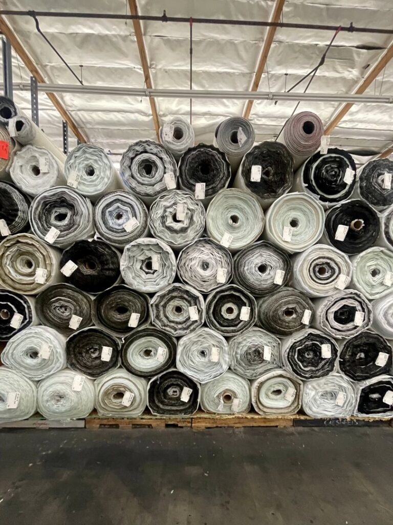 A large stack of carpet rolls, predominantly in dark colors, stored in the warehouse of Sy's Discount Carpets in Los Angeles, CA