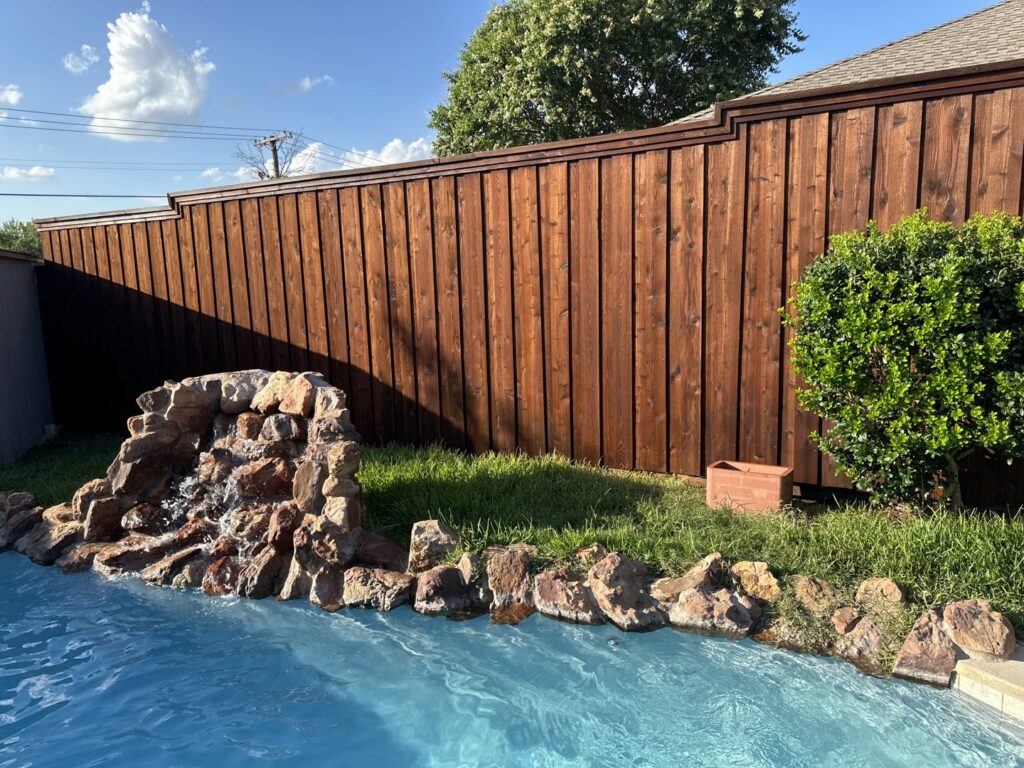 A stained wood fence providing a backdrop to a backyard pool with a decorative rock waterfall by Fence Defense, LLC in Plano, TX.