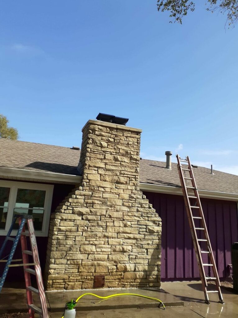 Ladders leaning against a house next to a tall stone chimney, indicating a chimney service job by A Step Above Chimney & Siding in Wichita, KS.