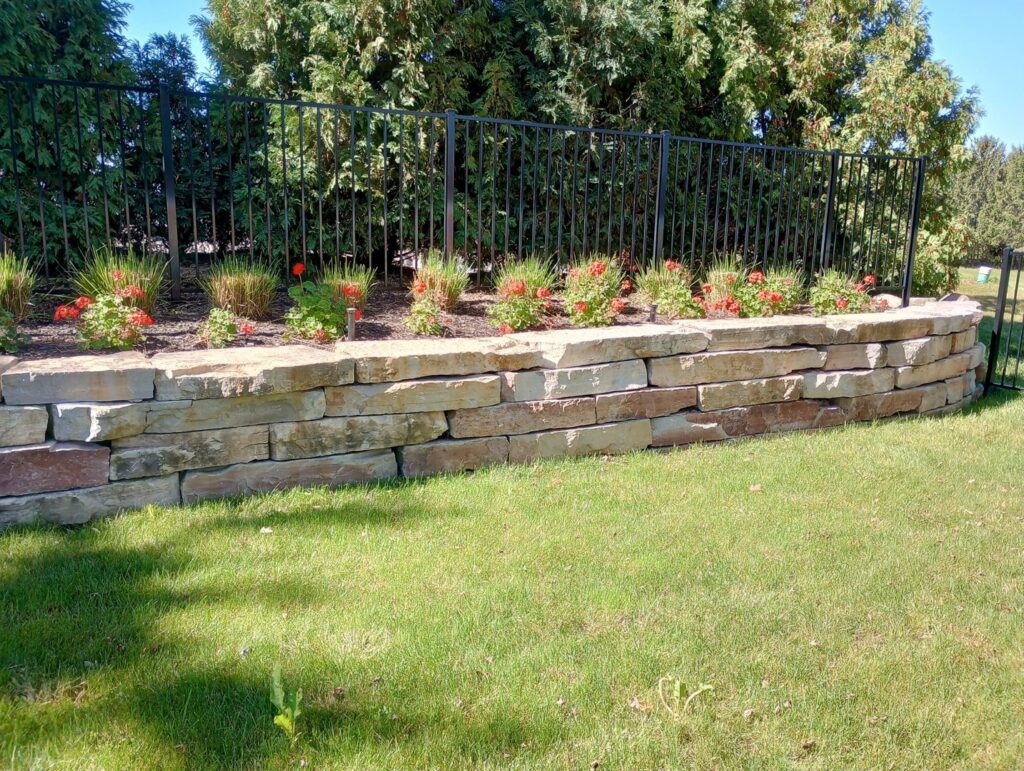 A decorative stone retaining wall with a vibrant flower bed next to a lush green lawn by Miguel Landscaping LLC in Janesville, WI.
