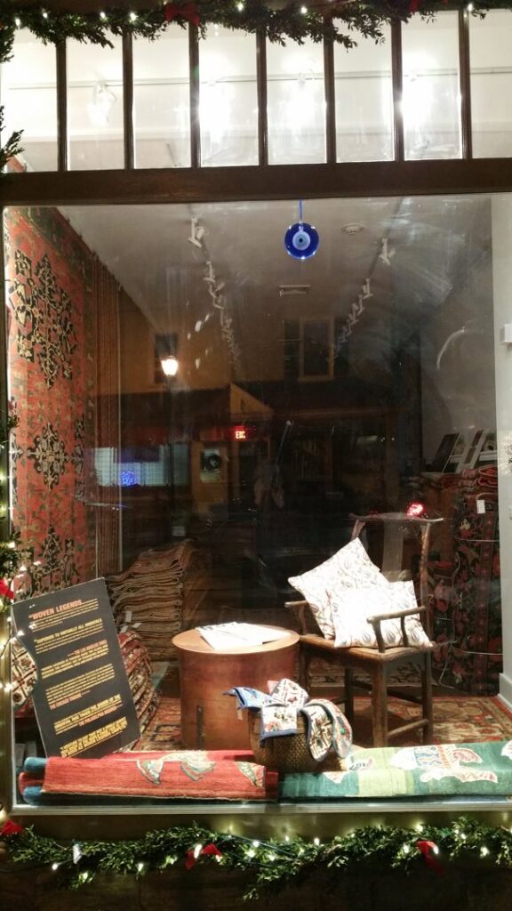 A festive storefront window display showcasing various rugs and home decor items from Woven Legends in Philadelphia, PA.
