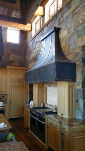 A tall, custom dark metal range hood with a curved design installed in a kitchen by Bensley Sheet Metal in Bozeman, MT.