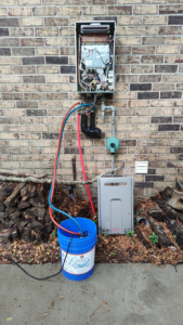 An outdoor tankless water heater being serviced and flushed with hoses connected to a bucket by CMC Mechanical in Goose Creek, SC.