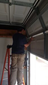 A technician on a ladder adjusting or repairing an upper garage door panel and track for Greeley garage door repair in Greeley, CO.
