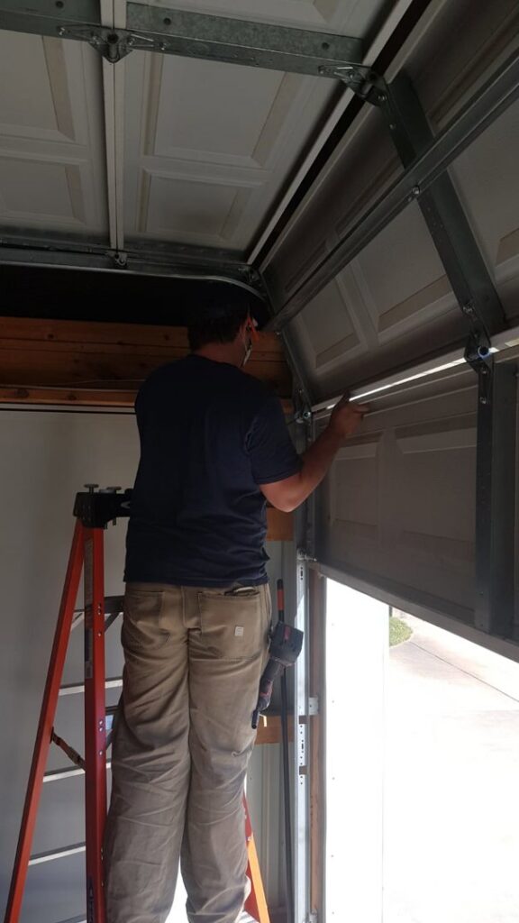 A technician on a ladder adjusting or repairing an upper garage door panel and track for Greeley garage door repair in Greeley, CO.