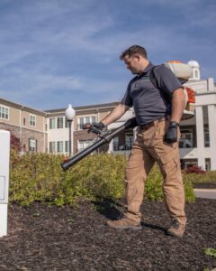 A Pest-Ops technician applying pest treatment with a backpack sprayer around a building in Knoxville, TN.