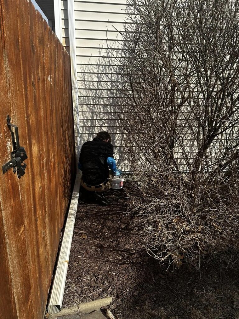 A Perimeter Pest Control DSM technician applying pest treatment in a tight space between a fence and a house in Des Moines, IA.