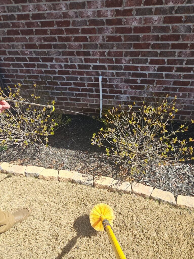 A technician from A Plus Pest Control using a long brush to treat or clean near a foundation in Huntsville, AL.