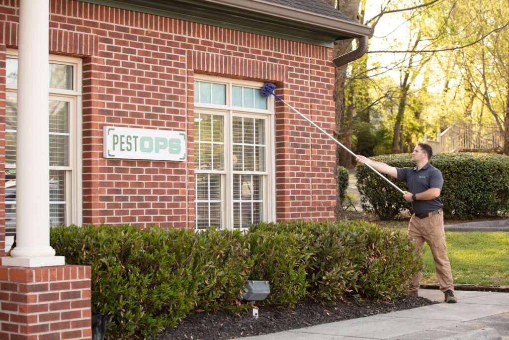 A Pest-Ops technician cleaning the exterior of a brick building with a long-handled brush for pest control in Knoxville, TN.