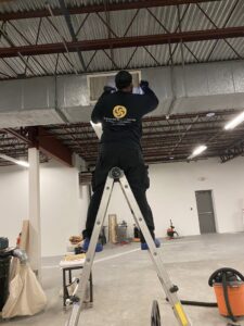 A technician from Boston Air Ducts Cleaning on a ladder, cleaning a ceiling air vent in Newton, MA.