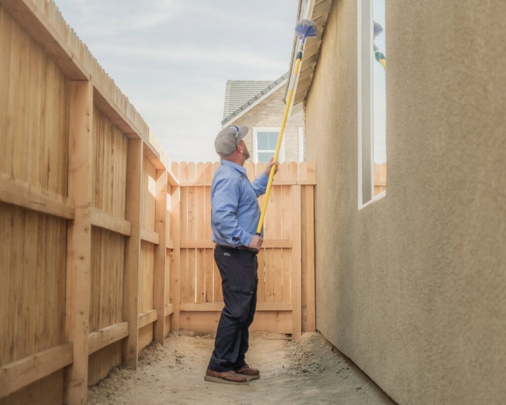 A Thrive Pest Pros technician cleaning cobwebs from the side of a house in a narrow passage in Clovis, CA.
