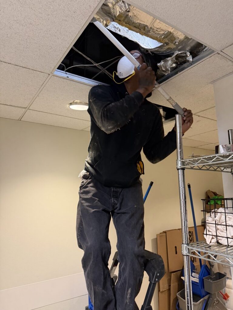 A technician from Boston Air Ducts Cleaning on a ladder, cleaning an overhead commercial air duct in Newton, MA.