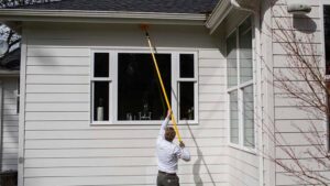 A pest control technician using a long pole to clean under the eaves of a white house for Pest Control Consultants in Sycamore, IL.