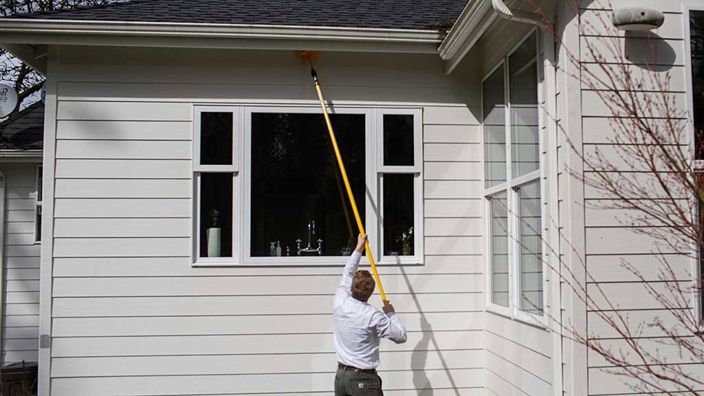 A pest control technician using a long pole to clean under the eaves of a white house for Pest Control Consultants in Sycamore, IL.
