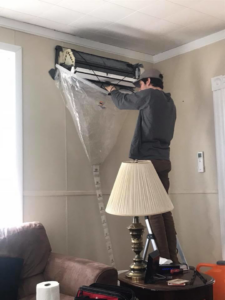 A technician on a ladder cleaning a mini-split heat pump unit with a protective cover at a client's home by Winks Heat Pump Cleanings in Bangor, ME